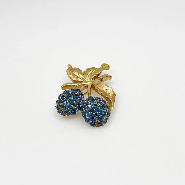 This vintage Crown Trifari brooch features a design of two berries suspended from intricate leaves and stems. The berries boast a captivating medium blue hue, beautifully set in a crafted gold-tone setting that gives the appearance of an invisible or pave setting