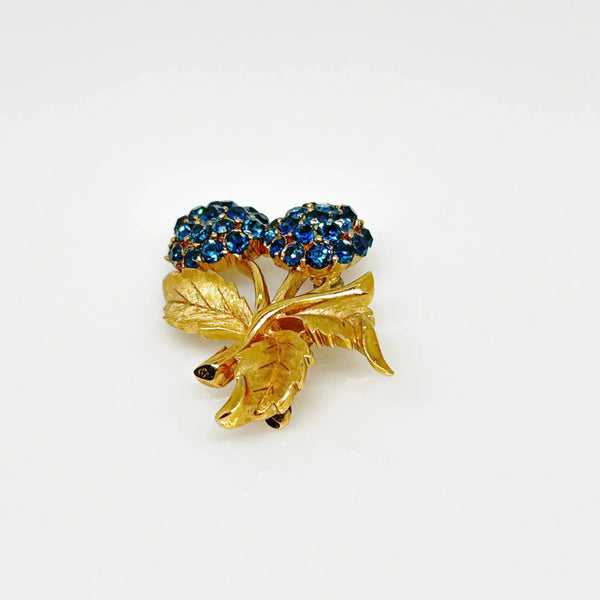 This vintage Crown Trifari brooch features a design of two berries suspended from intricate leaves and stems. The berries boast a captivating medium blue hue, beautifully set in a crafted gold-tone setting that gives the appearance of an invisible or pave setting