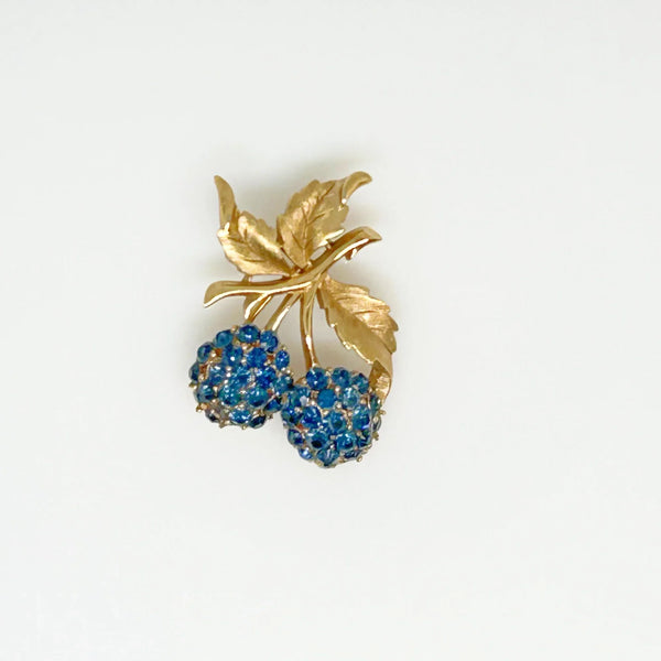 This vintage Crown Trifari brooch features a design of two berries suspended from intricate leaves and stems. The berries boast a captivating medium blue hue, beautifully set in a crafted gold-tone setting that gives the appearance of an invisible or pave setting