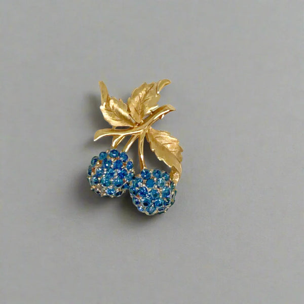 This vintage Crown Trifari brooch features a design of&nbsp;cluster of two blueberry-like fruit,&nbsp; adorned with blue faceted rhinestones, attached to a gold-tone stem with textured leave from intricate leaves and stems. The berries boast a captivating medium blue hue,
