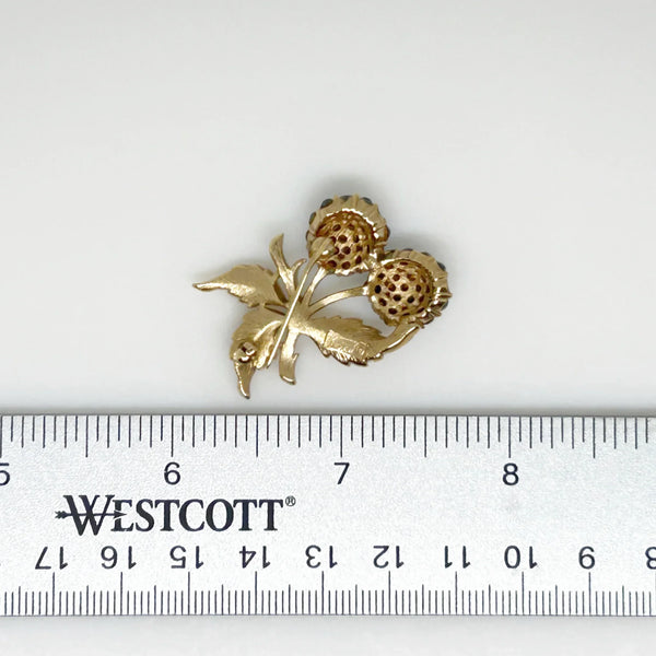 This vintage Crown Trifari brooch features a design of two berries suspended from intricate leaves and stems. The berries boast a captivating medium blue hue, beautifully set in a crafted gold-tone setting that gives the appearance of an invisible or pave setting