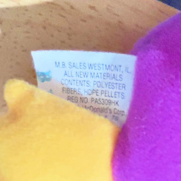 Close-up of a fabric tag sewn onto a plush toy, showing text including 'M.B.