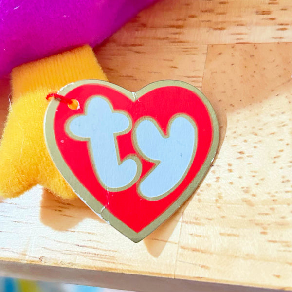 A heart-shaped Ty tag with a red face, light blue 'ty' lettering, and a metallic gold border, attached to a yellow plush toy on a light wood surface.