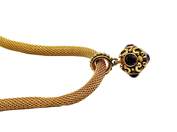 This 1990s pendant necklace features a meticulously crafted design with round purple faceted gemstones set in bezel-mounted antiqued gold metalwork. The ornamental pendant is suspended from a rounded mesh chain, measuring 16" secured with a toggle clasp.