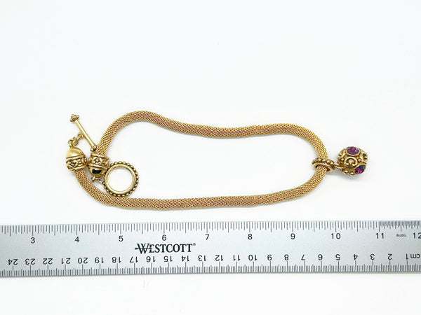 This 1990s pendant necklace features a meticulously crafted design with round purple faceted gemstones set in bezel-mounted antiqued gold metalwork. The ornamental pendant is suspended from a rounded mesh chain, measuring 16" secured with a toggle clasp.