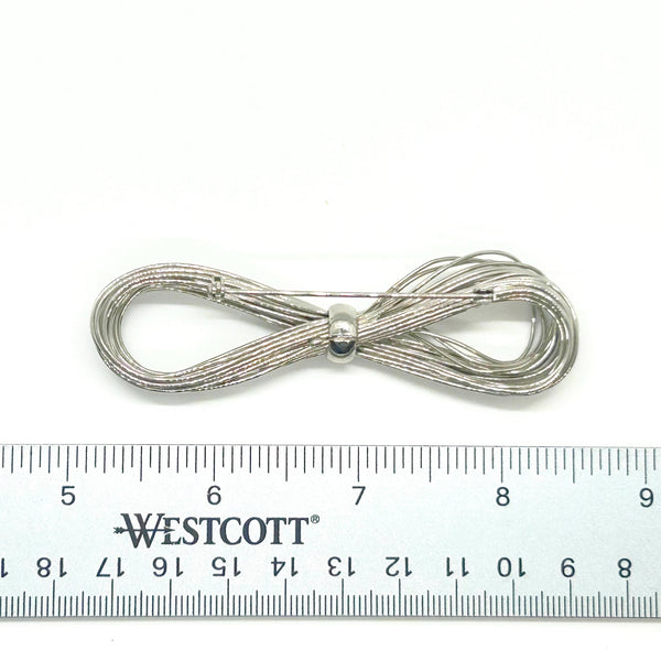 A silver-toned multi-strand wire bow brooch, featuring an infinity-shaped loop silhouette cinched with a smooth central band, displayed against a plain white background.
